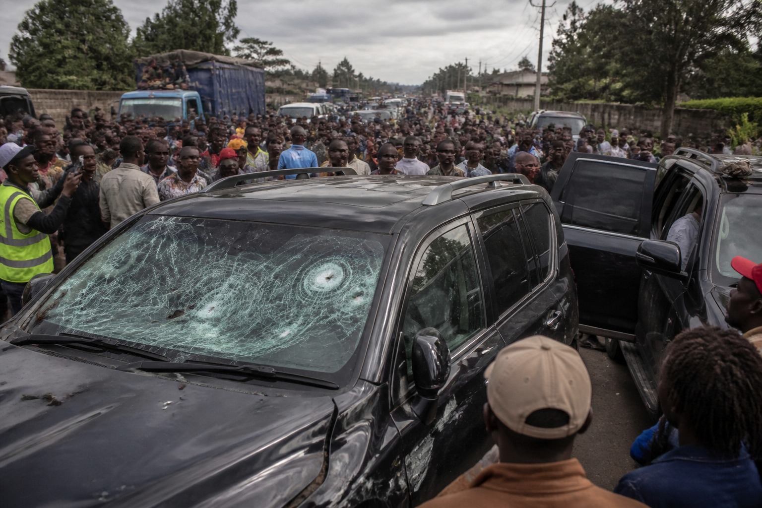 A black SUV with a shattered windshield, showing apparent bullet holes, is surrounded by a crowd of people on a busy road, consistent with a convoy attack.