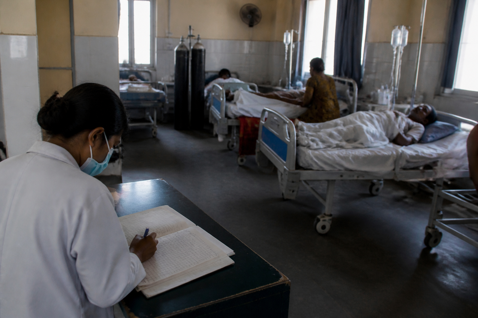 A medical professional records information in a ledger while patients rest in beds within a hospital ward, reflecting the challenges in data collection in healthcare networks.